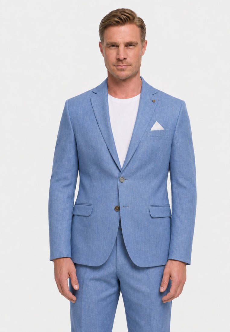 Homme portant une veste de costume bleu clair et un pantalon assorti, une chemise blanche en dessous et un mouchoir blanc dans la poche, debout devant un fond uni.