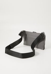 Borsa in nylon grigia con tracolla nera, dotata di un compartimento principale con zip e una tasca frontale con zip. Texture liscia, design piatto, dimensioni compatte.
