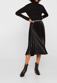 Černý rolákový svetr s knoflíkovými detaily na rukávech, kombinovaný s černou plisovanou midi sukní. Modelka má na nohou černé kotníkové boty.