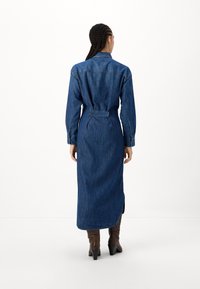 Robe chemise en denim, bleu foncé, à manches longues, taille ceinturée, fentes latérales, détail de col, portée avec des bottes à talons marron.