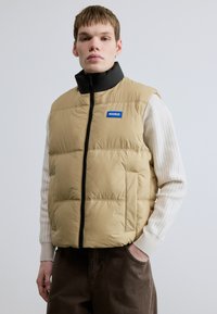 Beezhpaunane vest beeži värviga, kõrge musta krae, tõmblukuga sulgemine ja sinine "HUGO" logo. Omab tikandiga tekstuuri ja küljetaskuid.