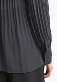 Czarna plisowana bluzka z długimi rękawami, zakończona mankietami na guziki oraz miękką, przejrzystą tkaniną. Dolny brzeg jest zaokrąglony, co nadaje całości zwiewny wygląd.