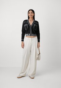 Cardigan nero corto con rifiniture bianche e tasche, indossato sopra pantaloni bianchi a vita alta. La modella tiene una piccola borsa a mano bianca.