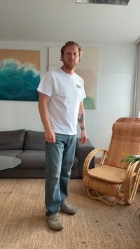 Weißes Baumwoll-T-Shirt mit grünem Levi's Logo, kombiniert mit blauen Jeans und grauen Clogs. Innenraum mit einem geflochtenen Stuhl und einer Couch.