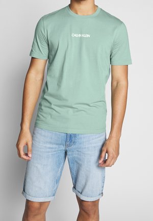 Homme portant un t-shirt Calvin Klein vert clair et un short en denim bleu clair retroussé, debout devant un fond gris uni.