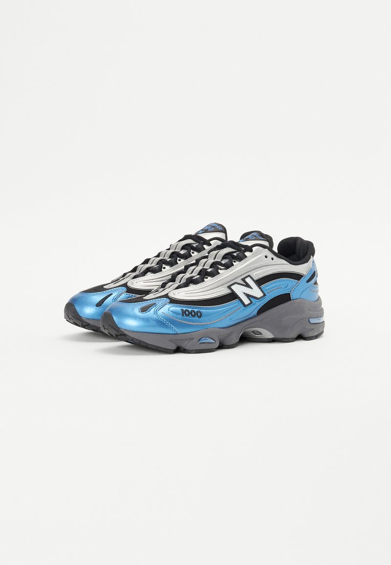 Blaue und silberne Sportschuhe mit einem Obermaterial aus Mesh und Synthetik, ausgestattet mit einem gepolsterten Kragen, schwarzen Schnürsenkeln und nummerierten Details an der Seite.