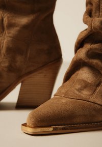 Καφέ καστόρινες μποτάκια αστραγάλου με σωρευτά ξύλινα τακούνια και χρυσή μεταλλική λεπτομέρεια με την ένδειξη "Bronx" στην εμπρόσθια άκρη της σόλας.