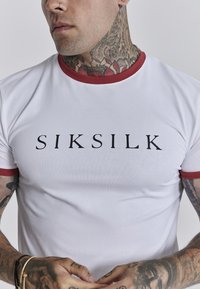 Witte T-shirt met korte mouwen en een rode geribde kraag, voorzien van een groot zwart "SIKSILK" logo. Gladde stof met een standaard pasvorm.