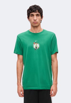 Młody mężczyzna w zielonej koszulce Boston Celtics z małym logo na piersi, stojący na jednolitym jasnym tle.