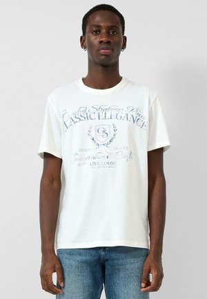 Jonge man draagt een wit T-shirt met blauwe vintage tekst en embleem, gecombineerd met een blauwe spijkerbroek, staand tegen een effen achtergrond.