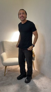 Hombre con camiseta negra y vaqueros oscuros de pie en una habitación con moqueta, cerca de un sillón blanco con textura y una mesa pequeña redonda con decoración.