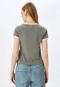 Olijfgroen t-shirt met korte mouwen en een ronde halslijn met witte rand, gemaakt van zachte stof, van de achterkant gepresenteerd met blauwe spijkerbroek.