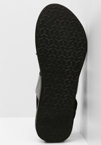 Musta kumisaapasoles, jossa on teksturoitu mehiläispesäkuvio. Varustettu kulmikkaalla reunalla ja kangasnilkoilla avoimelle muotoilulle.