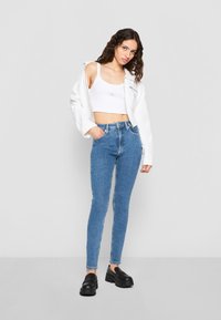 Skinny blue denim jeans with high waist and frontal pockets, paired with a white cropped tank top and a white oversized jacket. Black platform shoes.