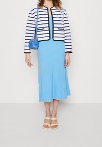 Kvinna i en blå midiklänning, vit och blårandig jacka, som bär en blå handväska, med beige klacksandaler mot en enfärgad bakgrund.