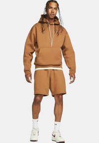 Felpa marrone con tasca a marsupio e lacci, abbinata a pantaloni corti coordinati, realizzata in tessuto morbido; indossata con scarpe da ginnastica bianche.