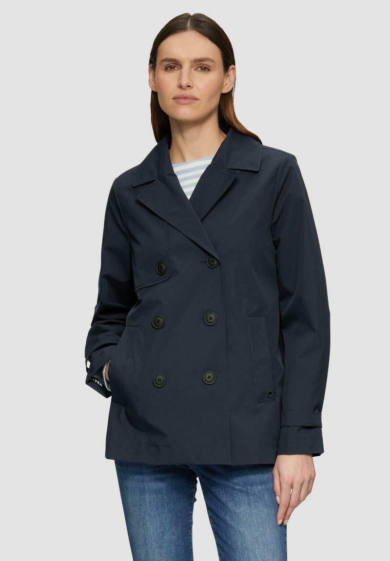 Manteau de marin bleu marine à double boutonnage avec de grands boutons noirs, poches latérales et col à revers, porté par-dessus une chemise rayée et un jean bleu.