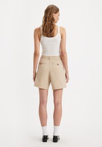 Shorts en coton beiges avec une coupe décontractée, dotés de poches arrière et de détails de couture subtils. Portés avec un débardeur blanc et des chaussettes.