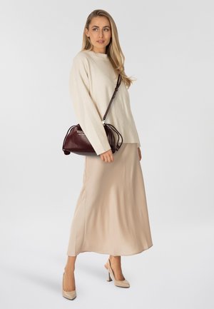 Pull en laine crème, jupe en satin beige et escarpins beiges. Sac à main bordeaux à la finition texturée et bandoulière tenu à la main.