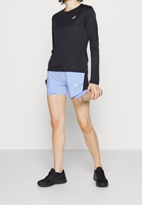 Svart långärmad träningströja, ljusblå shorts med meshpaneler i sidorna och svarta löparskor. Håller i en vattenflaska.