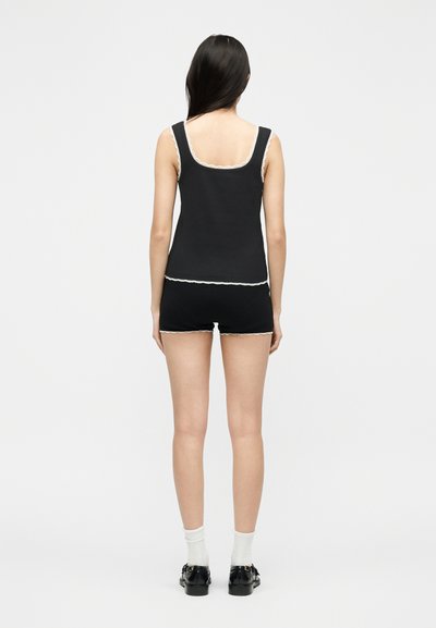 Femme vue de dos portant un débardeur noir et un short avec un bord en dentelle blanche, des chaussettes blanches et des chaussures noires sur un fond uni.