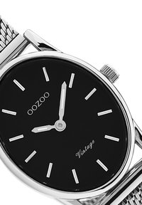 Montre à cadran noir avec aiguilles et index argentés, bracelet en maille en acier inoxydable et boîtier argenté. Présente les marques "OOZOO" et "Vintage".