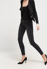 Jean skinny noir à cinq poches, en denim extensible. Porté avec un haut noir ajusté et des chaussures à talons hauts noires.