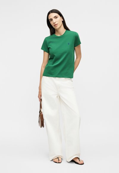 Γυναίκα που φοράει πράσινο κοντομάνικο μπλουζάκι, φαρδιά λευκή παντελόνα, μαύρα σαγιονάρες και κρατά μια μικρή καφέ τσάντα, στέκεται μπροστά σε απλό φόντο.