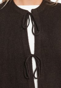 Mörkbrun stickad cardigan med rund halsringning, med två framknutar och en strukturerad yta. Tyget ser mjukt och varmt ut.