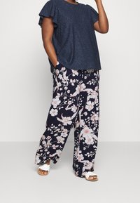 Vrouw in een marineblauwe short-sleeve top van oog genaaide stof, met brede bloemenprint broek en witte open slippers, met één hand in haar zak.