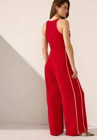 Röd ärmlös jumpsuit med vida ben, utrustad med rosa sidostripes, en bakre dragkedja och en figursydd överdel. Slät tyg och skräddarsydd design.