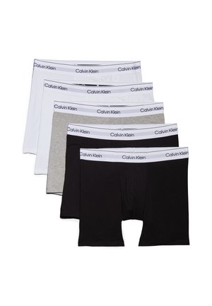 Atletisk mand iført hvide Calvin Klein boxershorts står barfodet med hænderne bag ryggen og kigger til siden mod en ensfarvet grå baggrund.