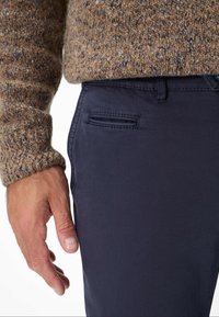 Marineblaue Hose mit glatter Textur und einer Fronttasche, kombiniert mit einem braunen, gesprenkelten Strickpullover. Hand ruht auf der Hose.