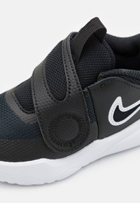 Zwarte sportieve schoen met een gestructureerde bovenkant, verstelbare haak-en-lussluiting, witte logo-accent en een gevoerde zool voor comfort.
