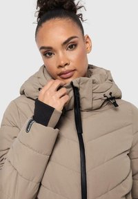 Beige gewatteerde jas met capuchon, voorzien van een geborduurd patroon, ritssluiting aan de voorkant en zwarte accentmanchetten. Close-upweergave van gezicht en hand.