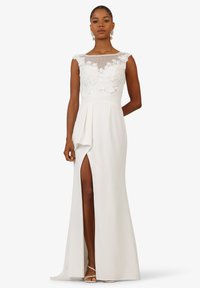 White gown with a lace bodice and sheer mesh neckline, featuring a high slit and draped detail on the skirt, smooth texture, and elegant silhouette.