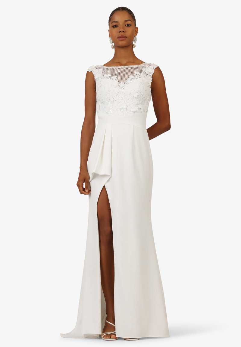 White gown with a lace bodice and sheer mesh neckline, featuring a high slit and draped detail on the skirt, smooth texture, and elegant silhouette.