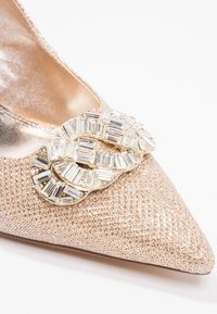 Kengät kärjellään, valmistettu metallinhohtoisesta vaaleanpunaisesta kankaasta, jossa on teksturoitu pinta. Edessä on kristallilla koristeltu solki.