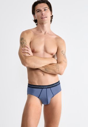 Mężczyzna bez koszulki z tatuażami na ramionach, ubrany w niebieskie majtki marki Sloggi, stojący z założonymi rękami, patrzący lekko w górę na jednolitym tle.