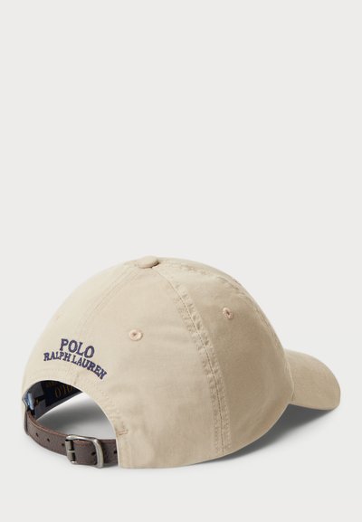Bēšas kokvilnas beisbola cepure ar izliekto malu, brūnu regulējamu siksniņu un tumši zilu izšūtu "POLO RALPH LAUREN" logotipu aizmugurē.