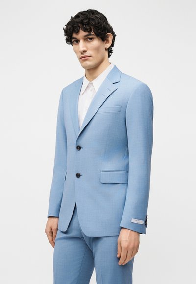 Νεαρός με σγουρά μαλλιά, ντυμένος με ανοιχτό μπλε κοστούμι δύο κουμπιών και λευκό πουκάμισο, στέκεται μπροστά σε απλό φόντο.