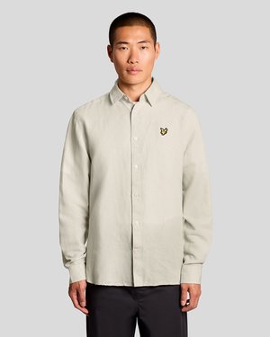 Bărbat purtând o cămașă cu nasturi de culoare bej deschis, cu un logo galben în formă de pasăre pe piept, stând în picioare pe un fundal gri simplu.