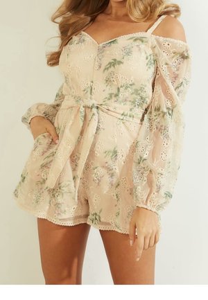 Vrouw in een beige bloemenkant romper met cold shoulder ontwerp, lange doorzichtige mouwen en een strikceintuur, poserend met haar hand in de zak.