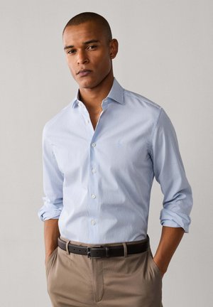 Hombre con camisa de botones azul claro a rayas con las mangas remangadas y pantalones beige con cinturón negro, de pie con las manos en los bolsillos.