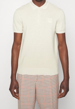 Mand iført en kortærmet, off-white polo-shirt med strikket tekstur og ribbet kant, kombineret med ternede bukser i beige, sort og rød.