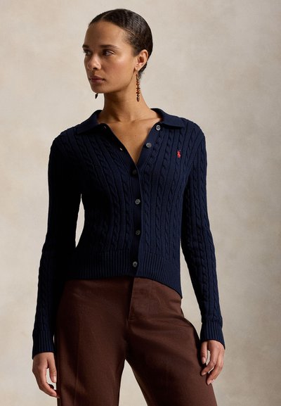 Nő tengerészkék, gombos, kötött kardigánban és barna nadrágban, oldalra nézve, semleges háttér előtt.