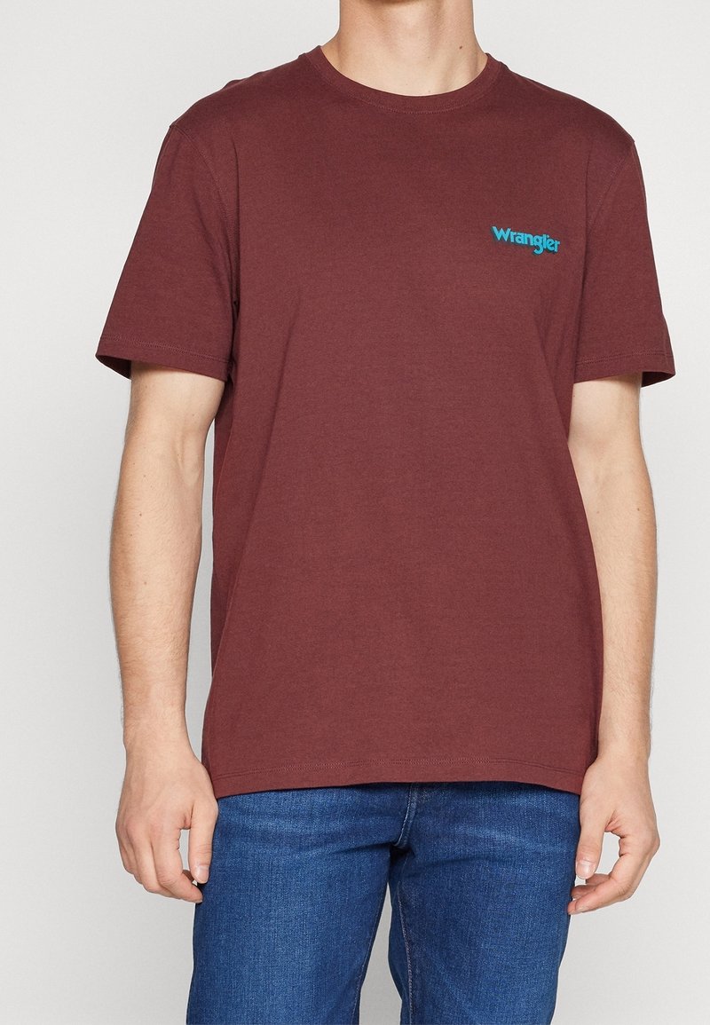 Homme portant un T-shirt Wrangler bordeaux à manches courtes et un jean bleu, debout les bras détendus le long du corps.