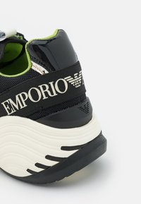 Svartvit sneaker med texturerad mesh-ovandel, glansiga detaljer och grön foder. Har en tydlig "EMPORIO ARMANI"-logotyp på hälen.
