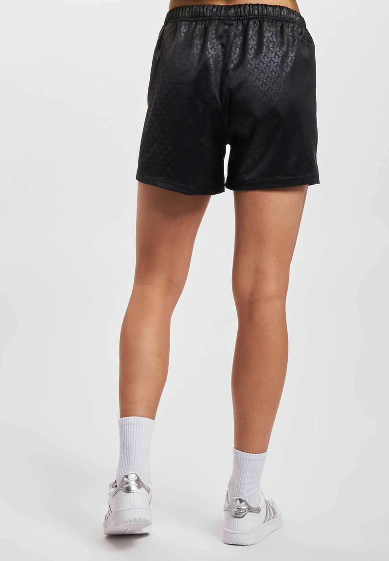 Personne portant un short noir texturé, des chaussettes blanches montantes et des baskets Adidas blanches, se tenant contre un fond clair uni.
