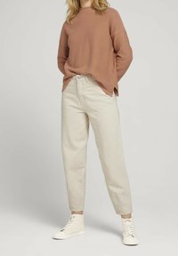 Beige Strickpullover mit rundem Ausschnitt, kombiniert mit hellbeigen, tapered Hosen und weißen High-Top-Sneakers. Weiche Textur überall.